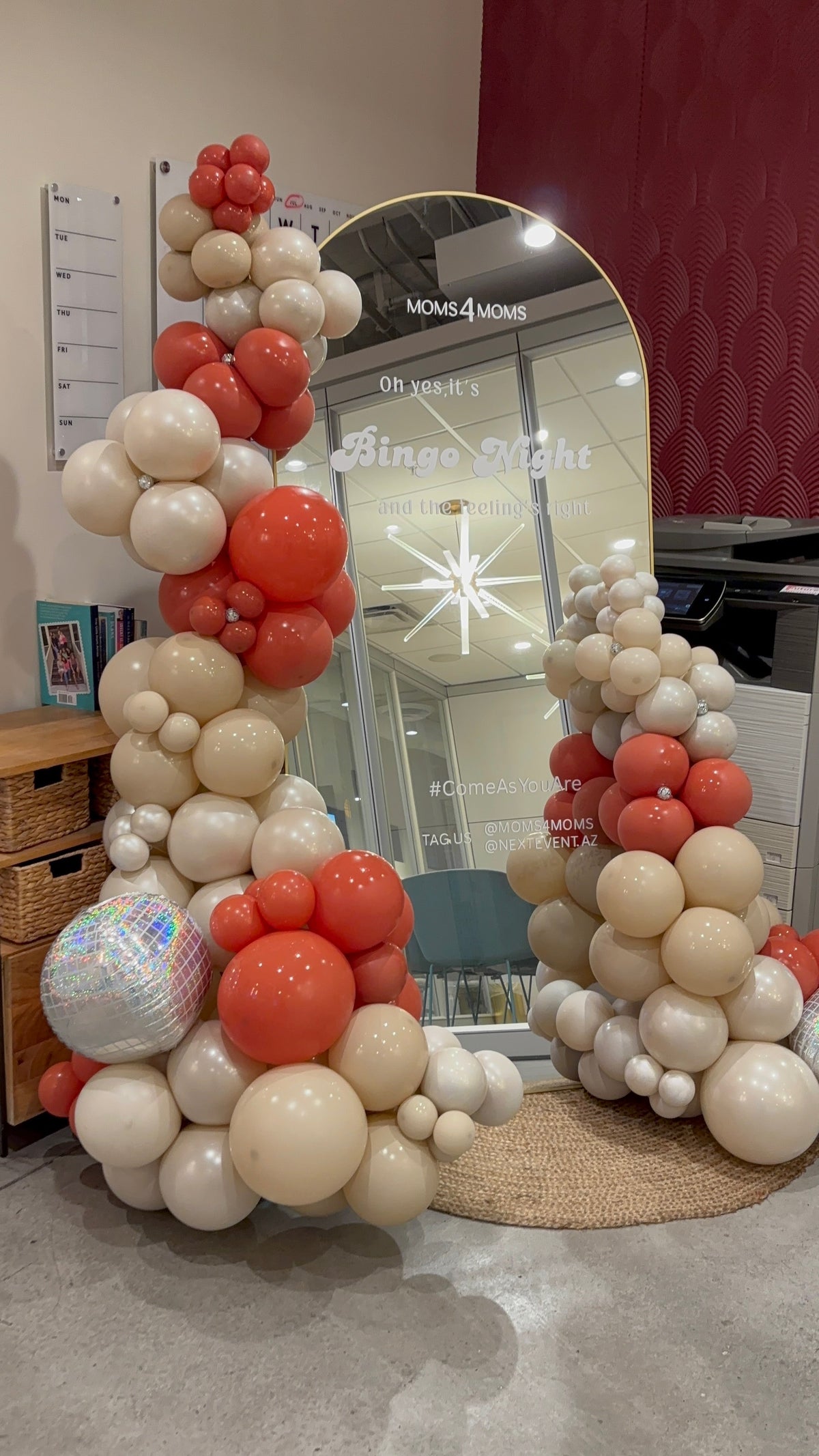 A decorative balloon garland installation with tall arches made of cream, burnt orange, and white balloons, plus a large disco ball balloon, set indoors in front of a gold arched mirror.